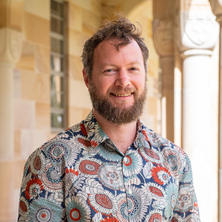 Dr Rowan Gollan standing in UQ Great Court with sandstone pillars wearing a brightly-patterned floral shirt.